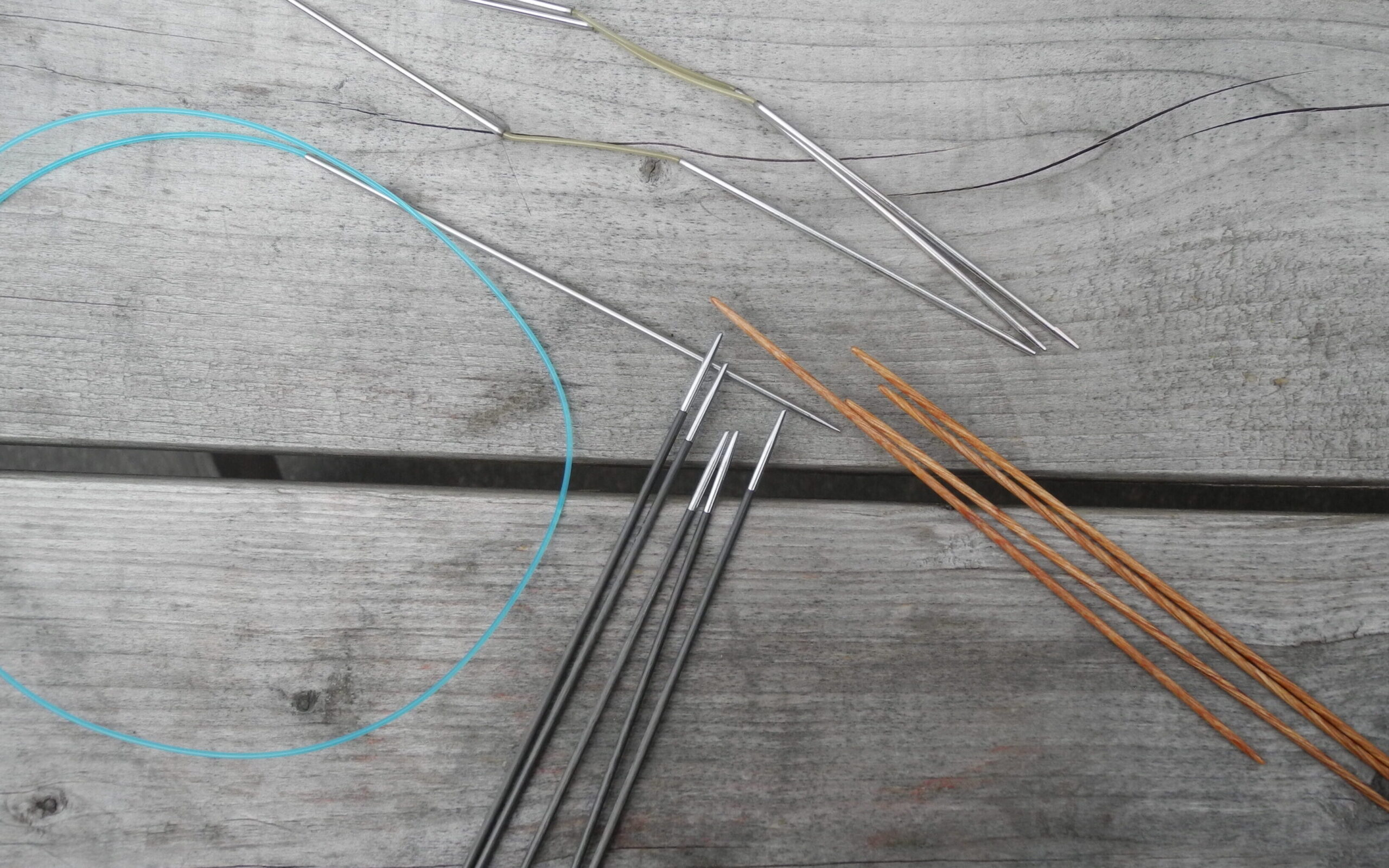 close up of different knitting needles including circular needles with blue cables metal and wood double pointed needles and flexiflips on wood background