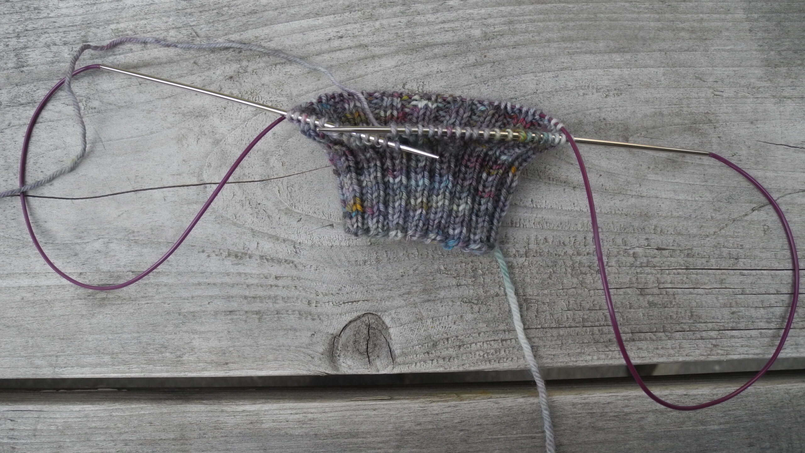 partially knit sock on long circular knitting needles with purple cables on wood background