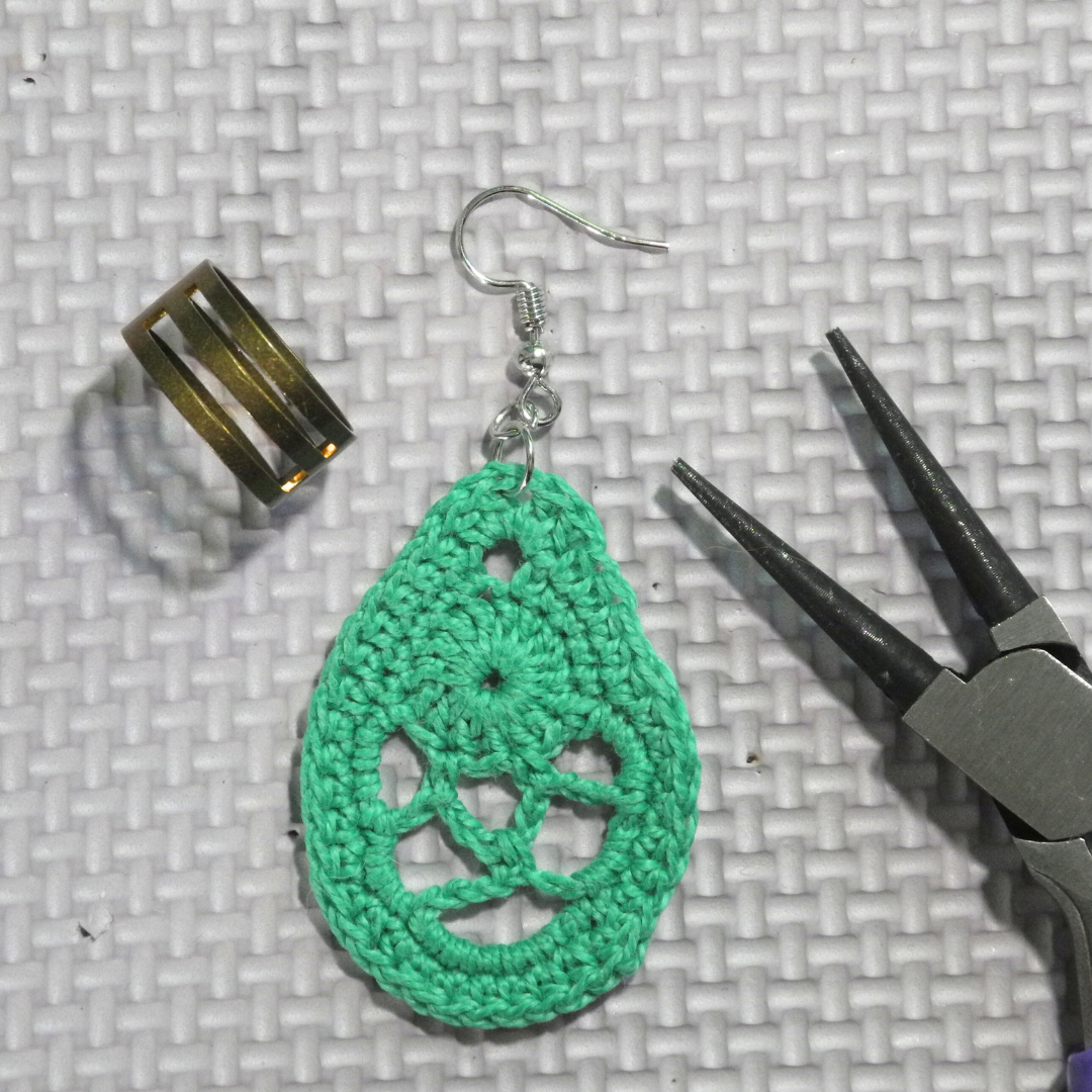 copper jump ring opener next to green crochet earring in dragon egg pattern with silver hook next to round nose jewelry pliers