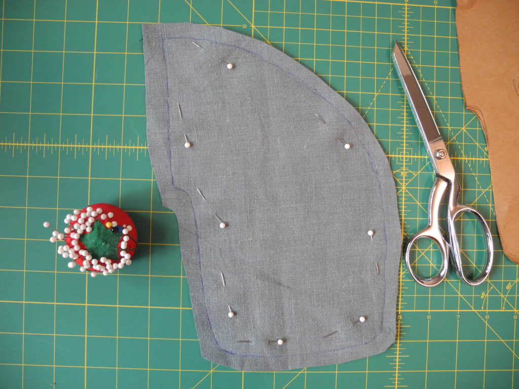 blue linen fabric cut into pocket shape with red pin cushion and silver scissors on green grid background 