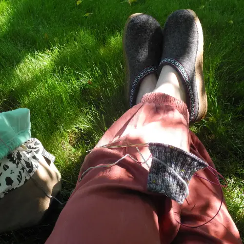 partially knit sock on lap. legs crossed and outstretched with orange pants and grey clog shoes on feet. legs are resting in sun dappled grass with teal, white, black and brown bag sitting next to the lap on the grass