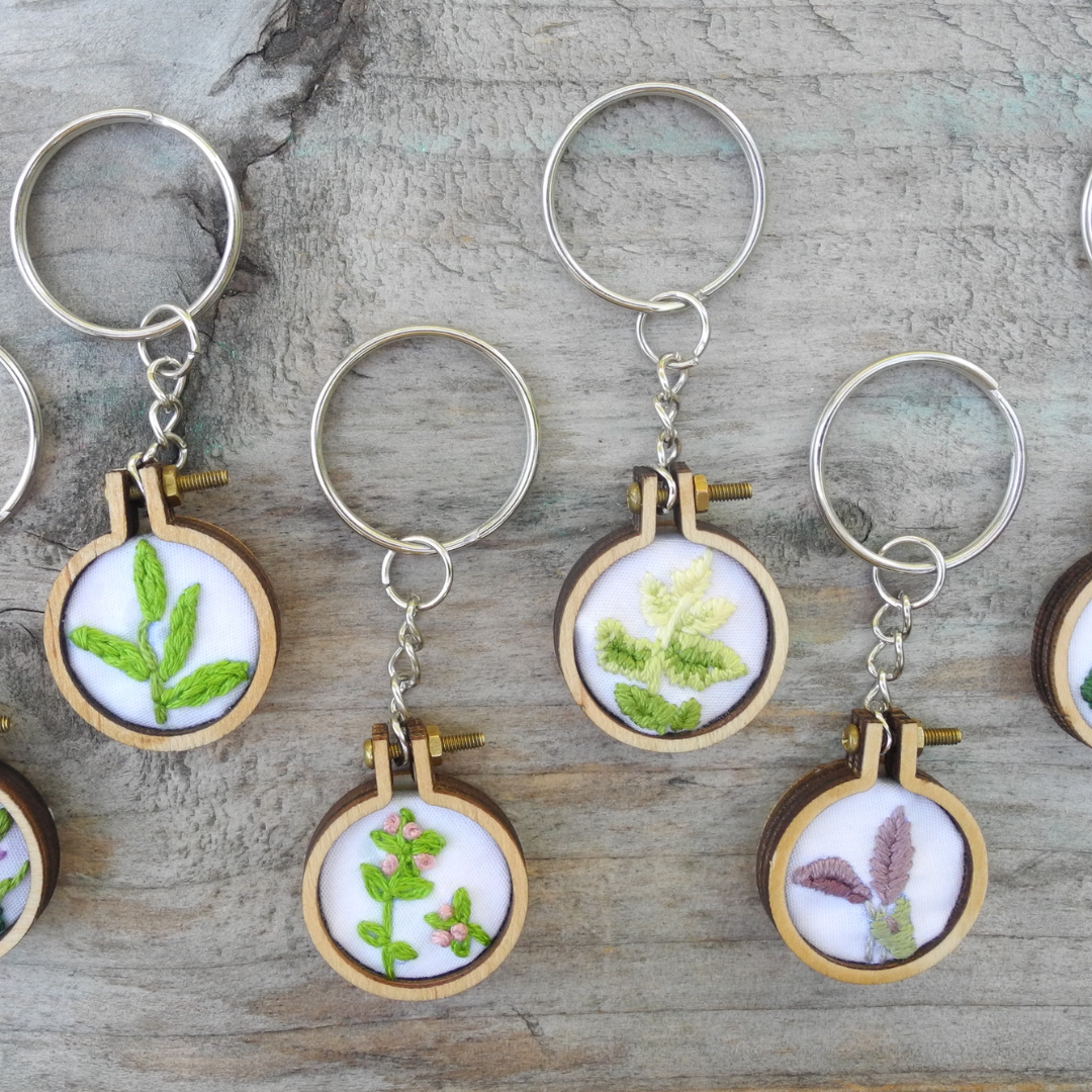 four keychains with embroidered herbs. left keyring has tarragon, middle left has thyme, middle right has oregano, right has sage embroidery