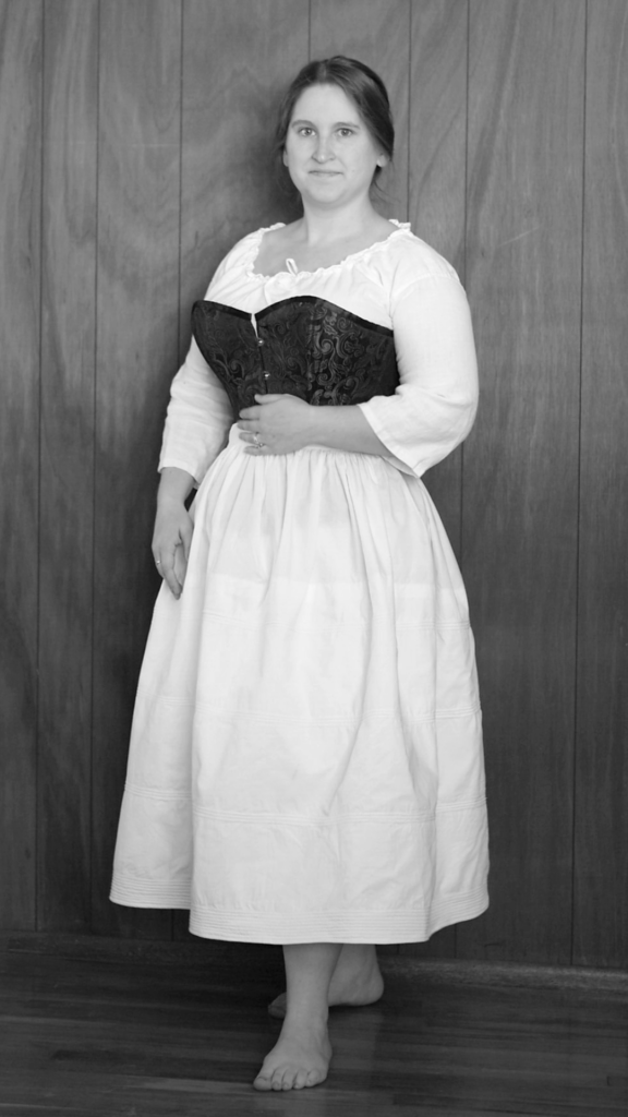 Oval black and white photo of a white woman standing in bare feet with a white chemise and black corset wearing a white corded petticoat 