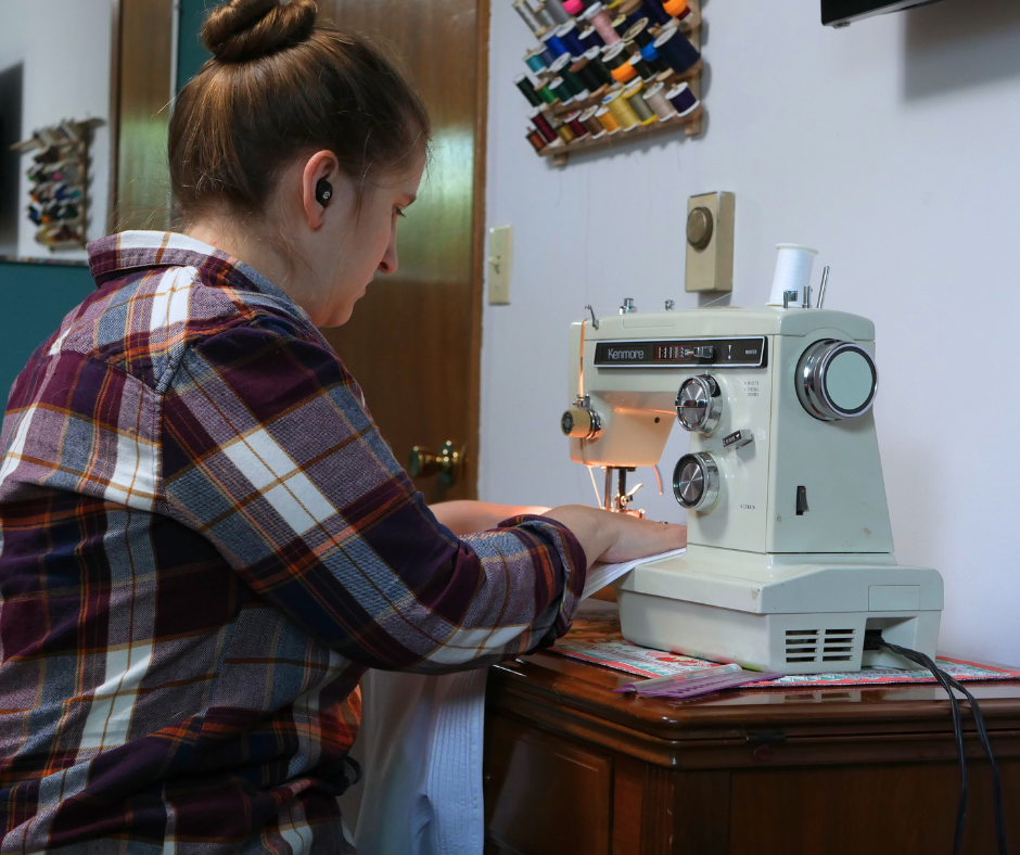 picture of a white woman wearing a maroon, white, and orange plaid shirt with her hair in a high bun sitting at a vintage kenmore sewing machine sewing cords into a corded petticoat. 