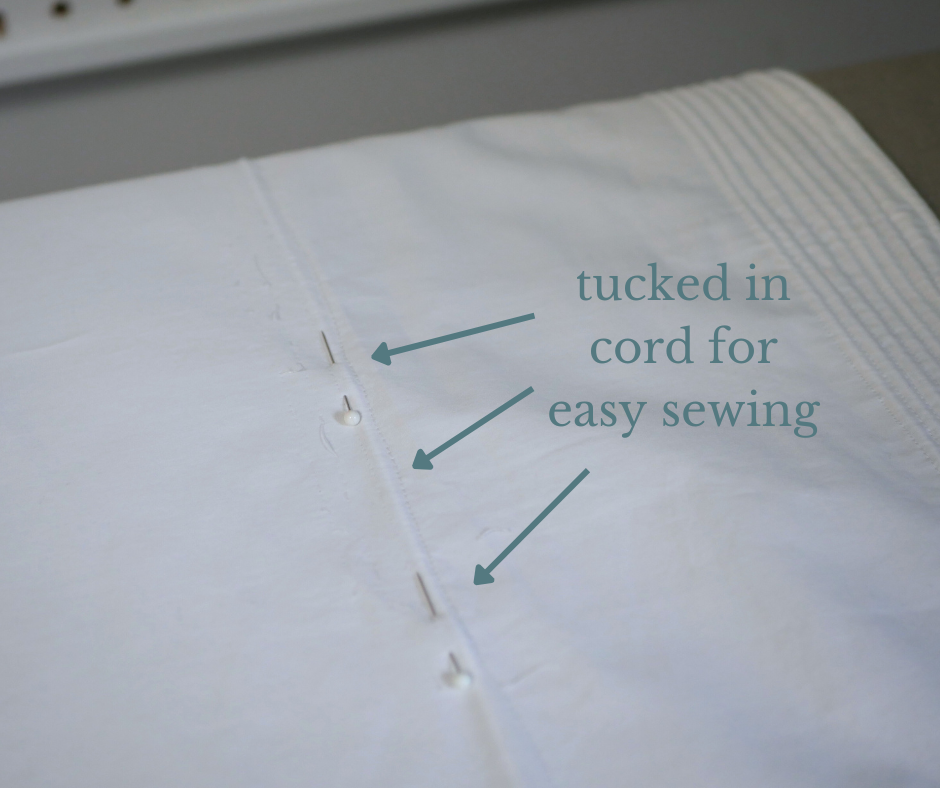 close up of a white corded petticoat with a row of cording about to be sew into place neatly nestled against a seam line, held in place by two straight pins. The words tucked in cord for easy sewing are written in teal font with three teal arrows pointing to the cord. 