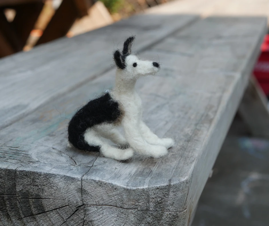 a black and white needle miniature needle felted dog in a sitting position on a wood bench. 