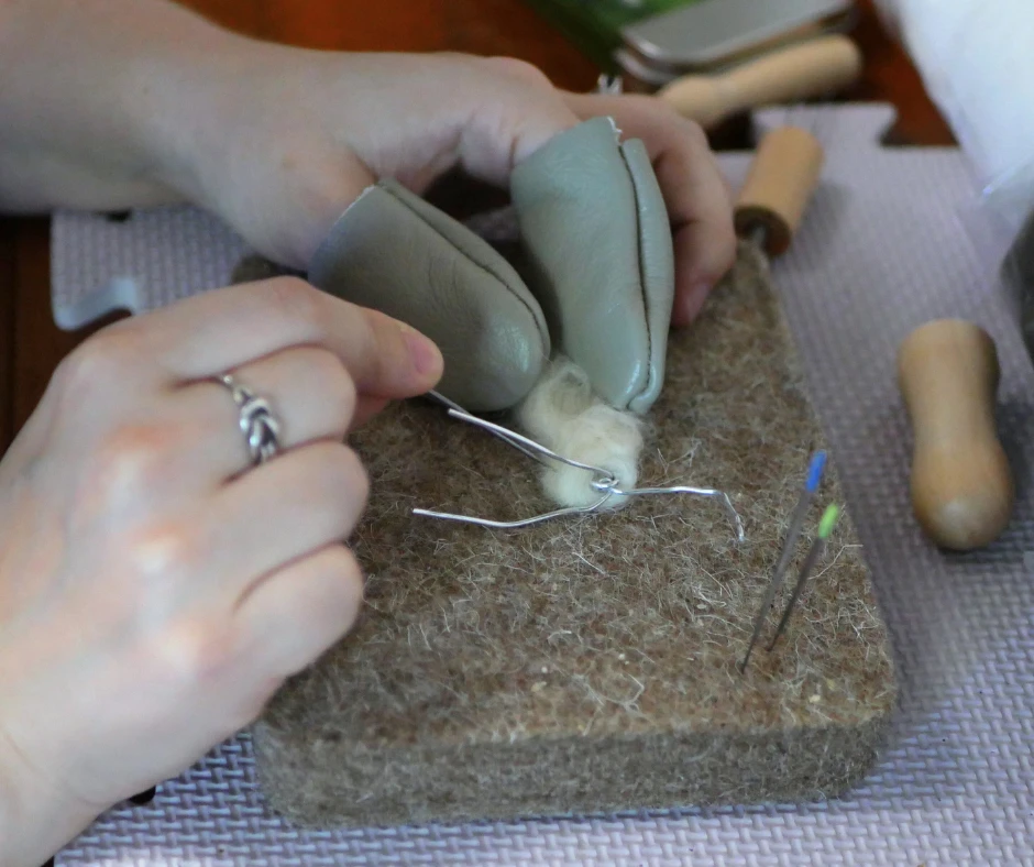 a pair of white hands work on needle felting some white wool onto a wire frame. The left hand has grey finger guards on the pointer finger and thumb. The right hand holds a felting needle. 