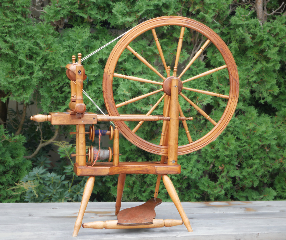 a light colored wood norwegain spinning wheel with double drive tension sitting on a grey wood surface with a greenery background. 