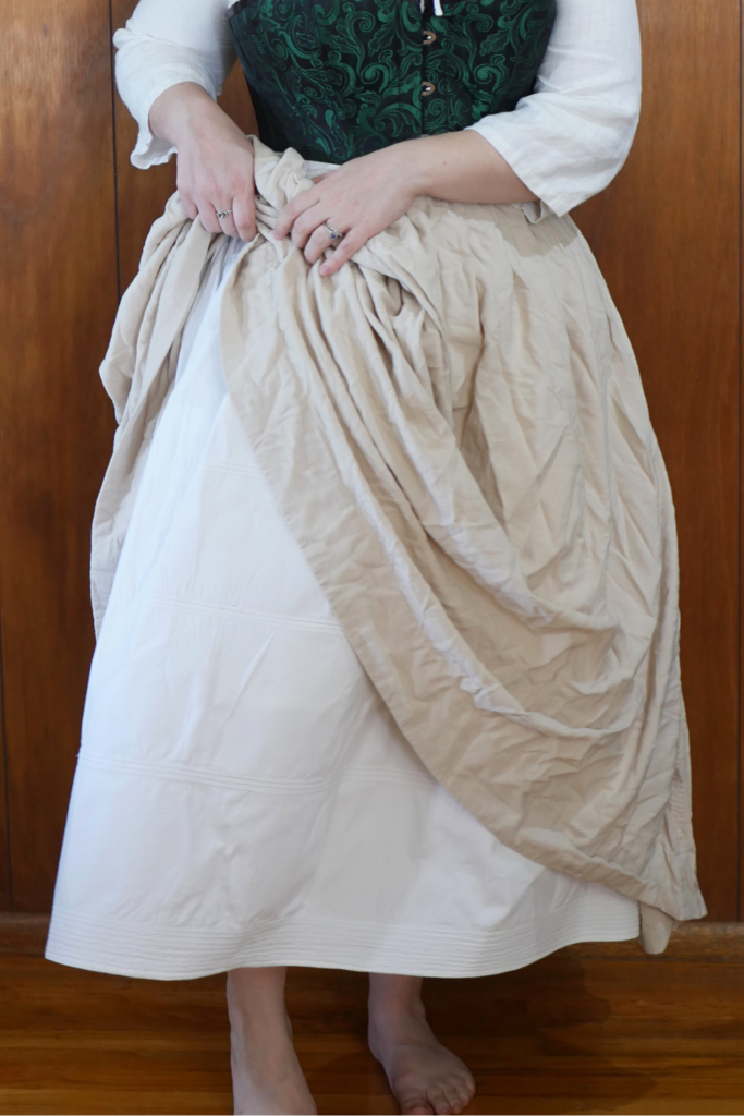 close up of a white woman wearing a starched white corded petticoat with an off white skirt over top being held up on one side to show the petticoat underneath. 
