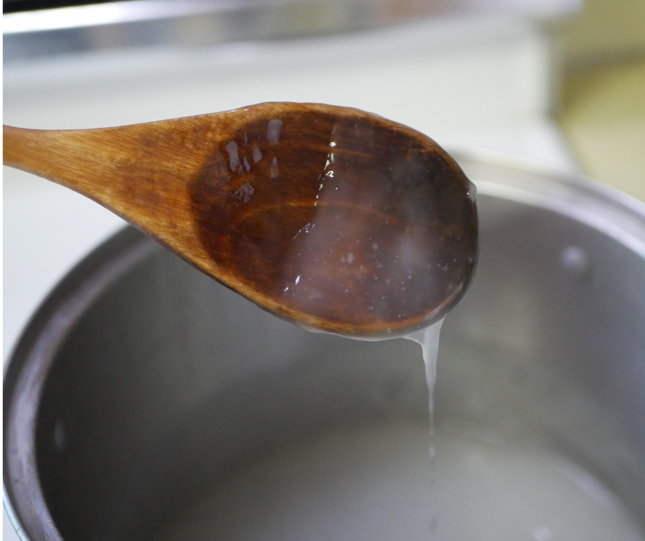 close up of a wooden spoon with clear viscous liquid dripping off of it into a steel pot. 