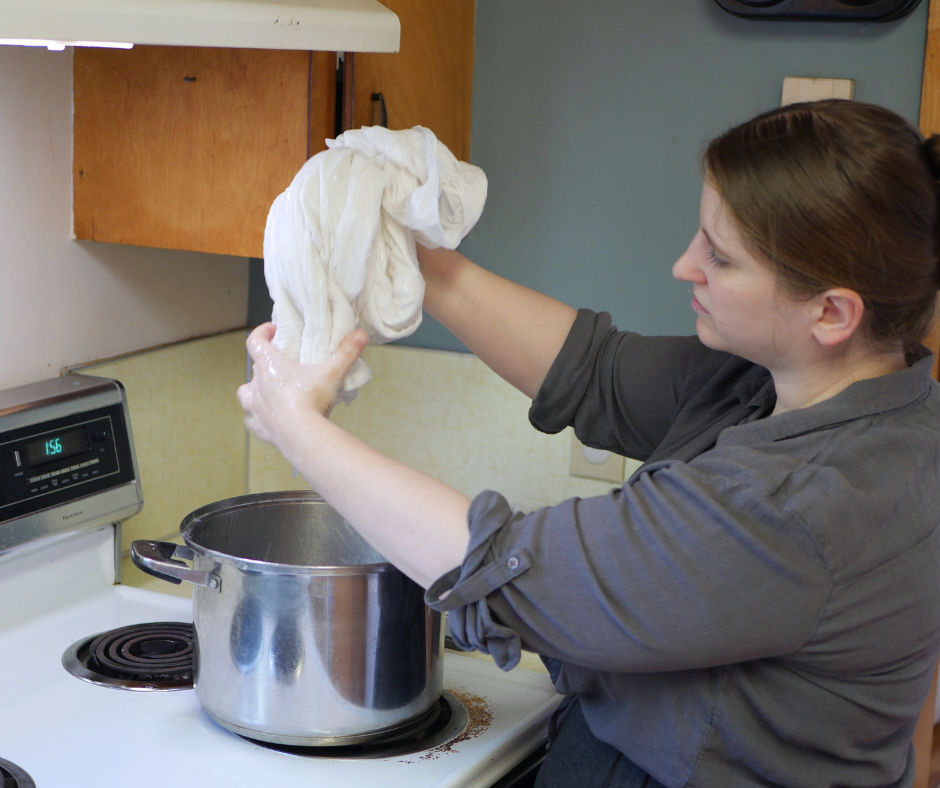 a white woman in a grey shirt wrings liquid laundry starch out of a white corded petticoat over a steel pot on a white stove top. 