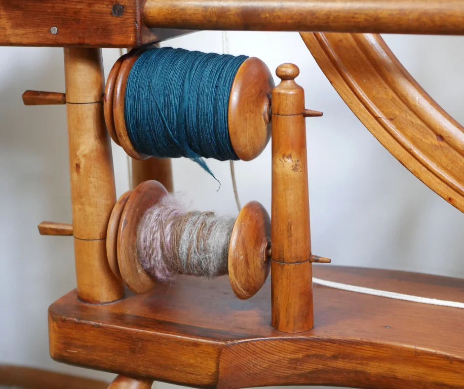 close up of a lazy kate on a spinning wheel with two bobbins. The bottom bobbin is partially filled with pink, brown and grey yarn. The top bobbin is completely full of teal yarn single ply. 
