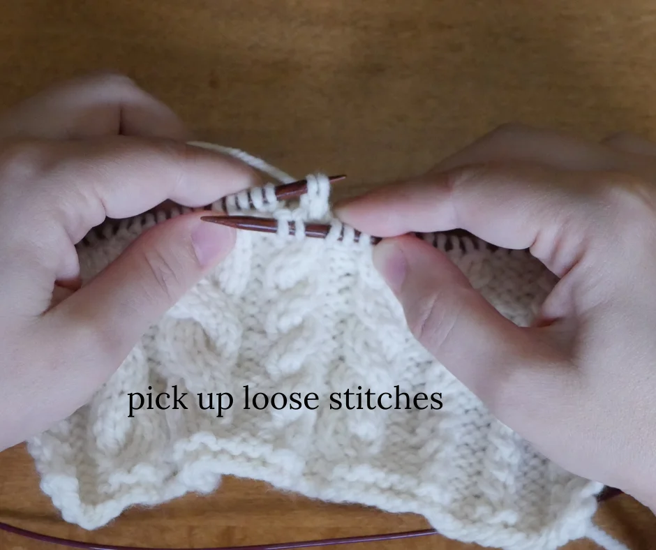 image of white knitted fabric on dark wood knitting needles with the left hand needle picking up two stitches for cable. Image labeled pick up loose stitches. 