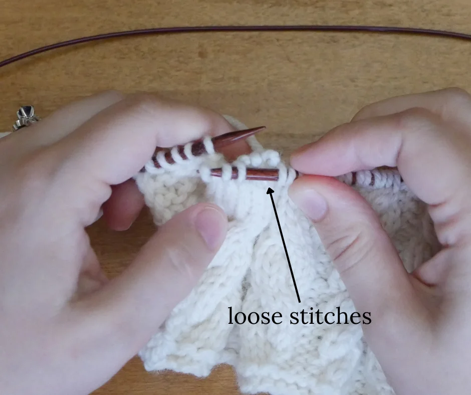 white knitted fabric on dark wood knitting needles with three loose stitches behind the right needle. Image labeled loose stitches. 