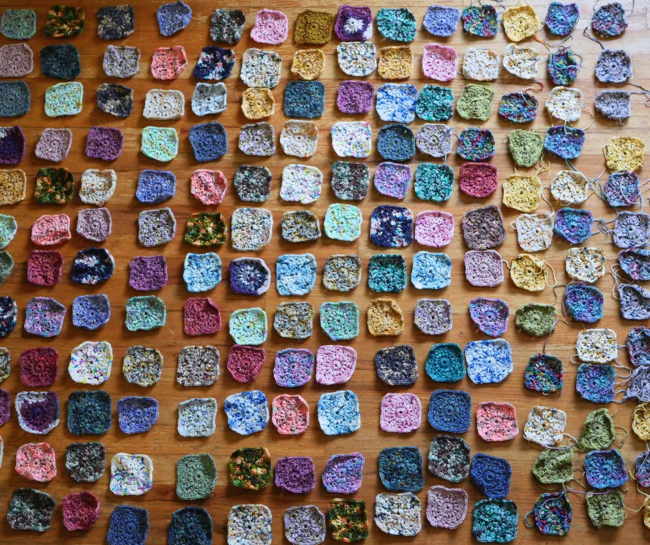a lot of crochet granny squares laid out on a wood floor in a grid pattern in a variety of colors. 