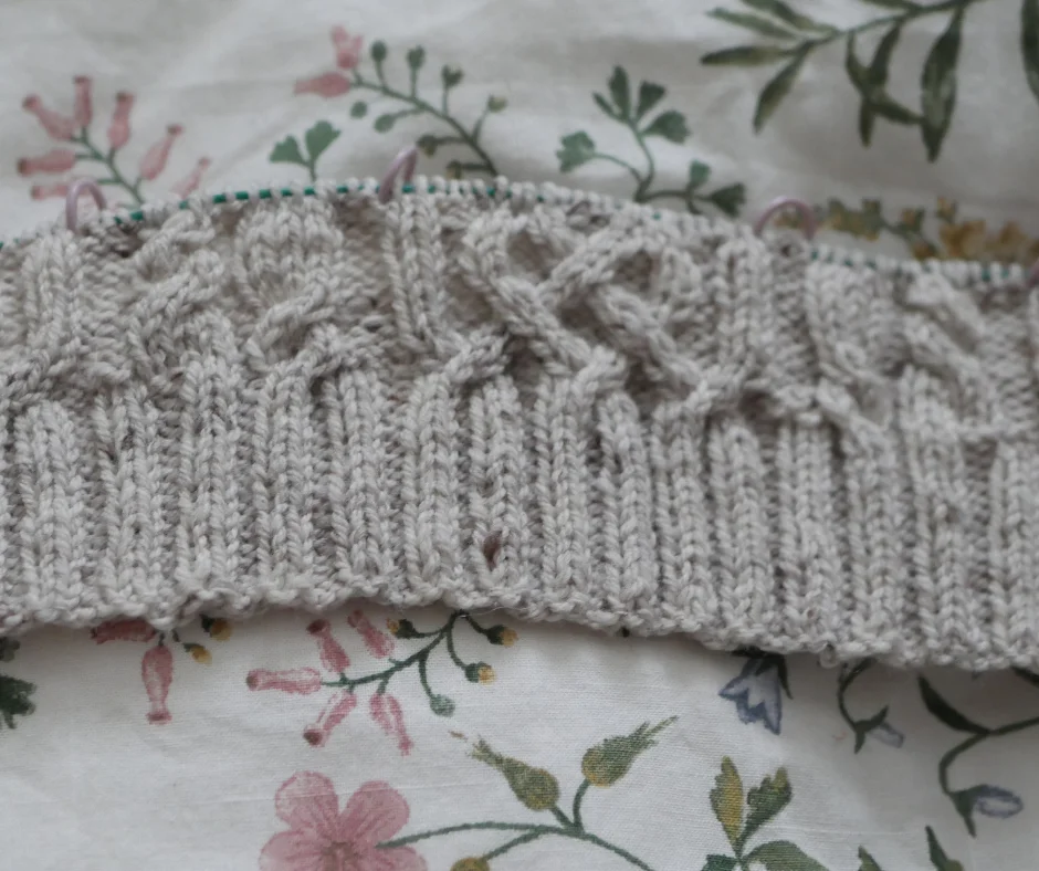 a closeup of grey wool knit into rows of rib stitch and a cable knit patter resting on a white cloth with floral pattern. 