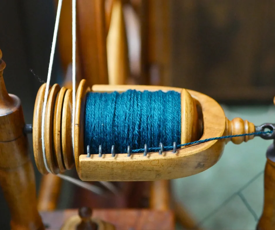 close up of the flyer of wood spinning wheel with a nearly full bobbin of three ply teal wool yarn. 