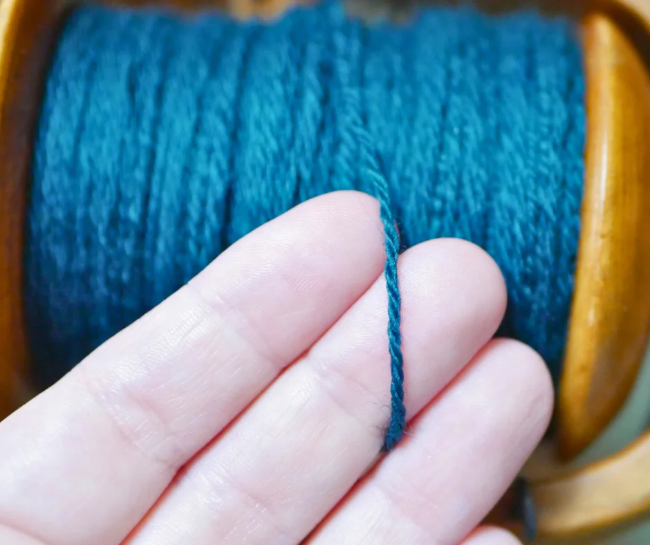 closeup of teal yarn laying across a white persons fingers to give an indication of the thickness of the yarn. 
