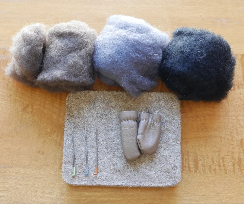 image of needle felting supplies. There are three colors of wool roving, brown, grey, and black, a wool mat, three different felting needles, and a pair of leather finger guards. 