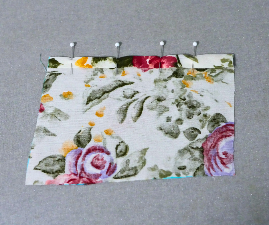 a square of floral fabric with one edge folded down and four straight pins with white balls on the top holding the folded fabric down. 