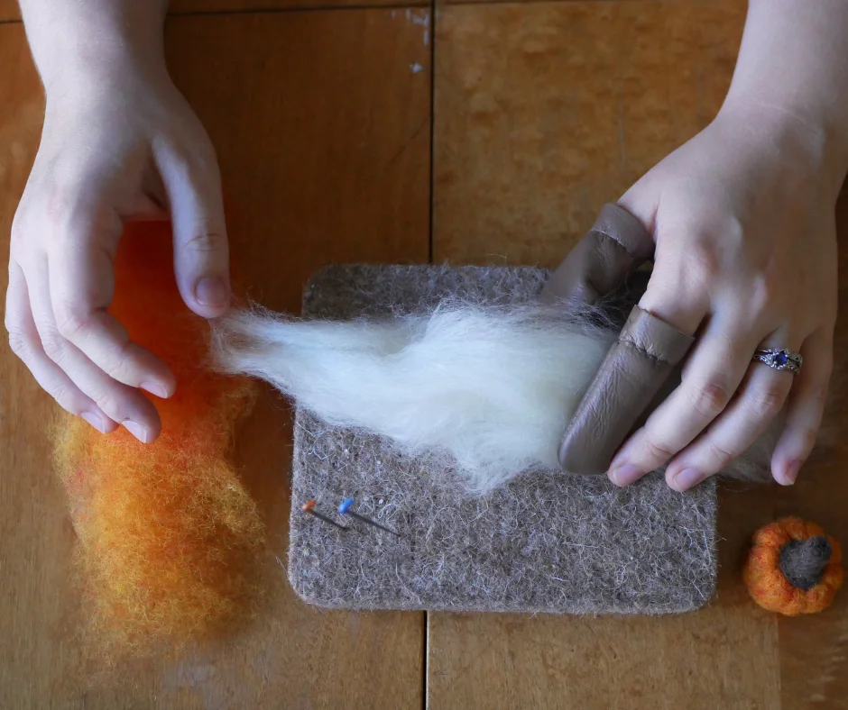 image of two white hands with leather finger guards on the pointer finger and thumb of the left hand resting on a wool mat with white wool in the left hand. Under the right hand is some orange wool roving. There are two felting needles in the wool mat and a small felted pumpkin in the lower right side of the image. 