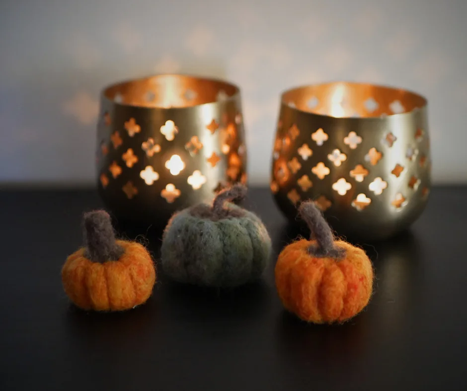 image shows a trio of mini needle felted pumpkins. The left and right pumpkins are orange, the center pumpkin is green. In the background are two brass candle holders with four sided flower cut outs and candlelight coming from inside. 