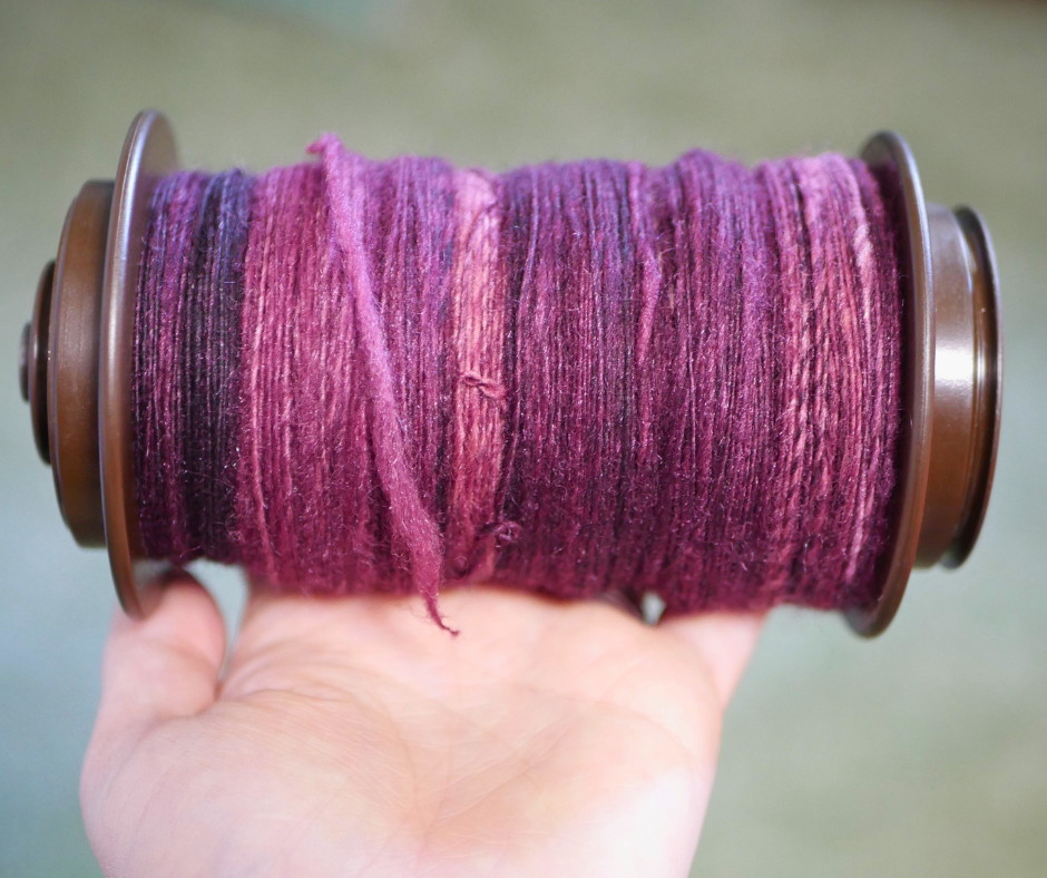 Image of a brown spinning wheel bobbing full of purple single ply yarn in various shades being held by a white person's hand. 
