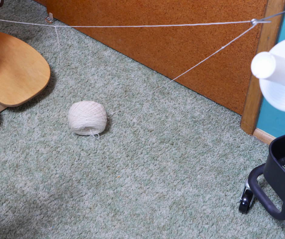 Image of a cake of white yarn that has fallen off of the ball winder onto the carpeted floor. 