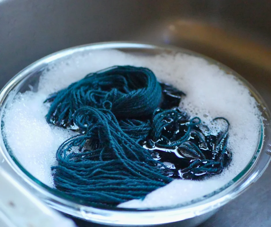 Image of a clear glass bowl in a metal sink full of teal yarn and bubbles. 