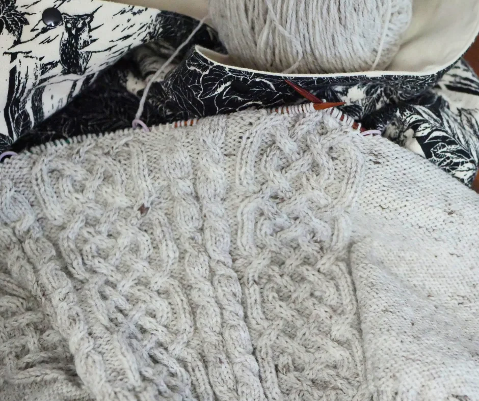 Image of a partially completed cream colored cable knit sweater on wood knitting needles resting on a black and white fabric bag.