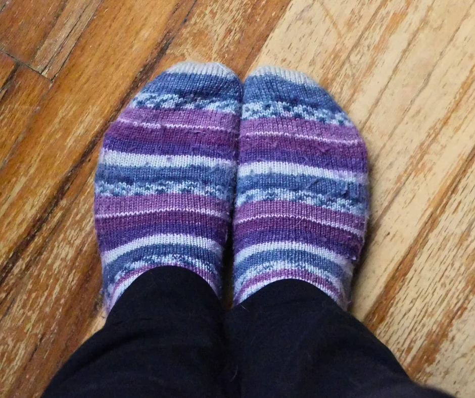 Image of a pair of feet wearing hand knit socks standing on a wood floor. Socks have purple, blue, and white stripes. 