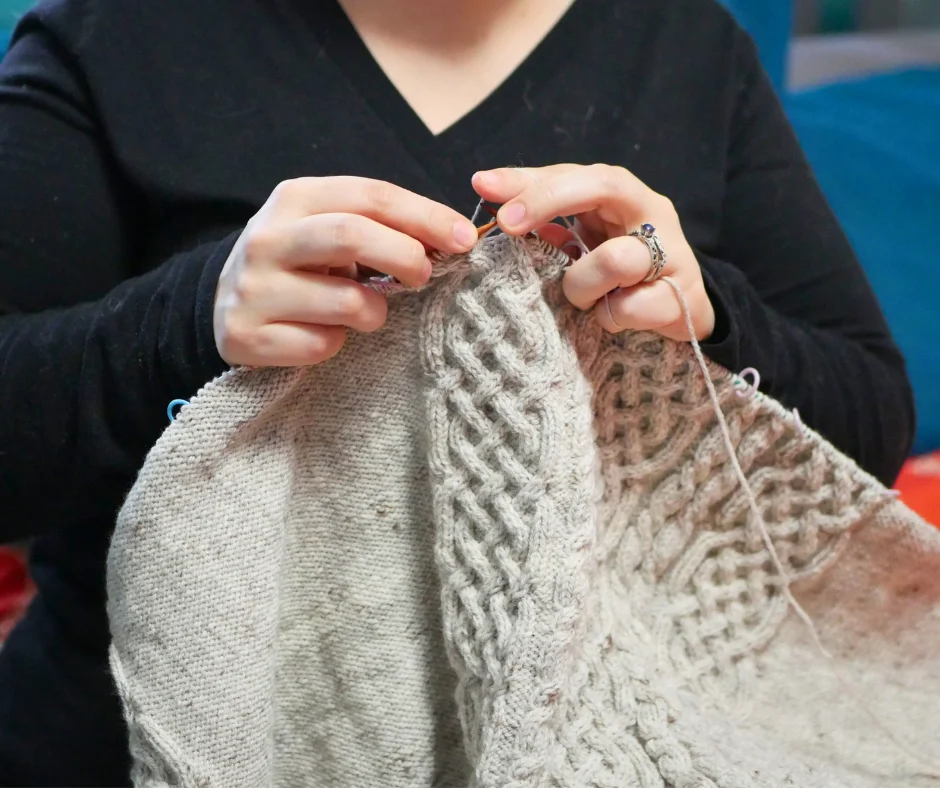 Image of a pair of white hands knitting on a cream colored fabric with a Celtic cable motif. 