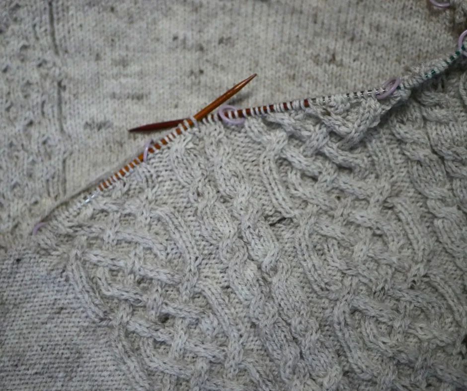 Image of a cream knit fabric still on knitting needles with a Celtic cable motif. 