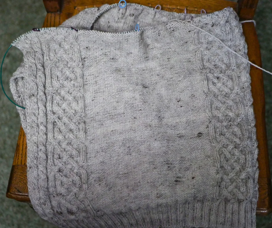 Image of a partially knit grey sweater with a Celtic cable motif resting on a wood chair. 