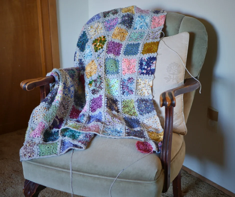 Image of a partially completed crochet granny square blanket resting over the back and one arm of a vintage style chair with sage green velvet upholstery and wood arms and legs. 
