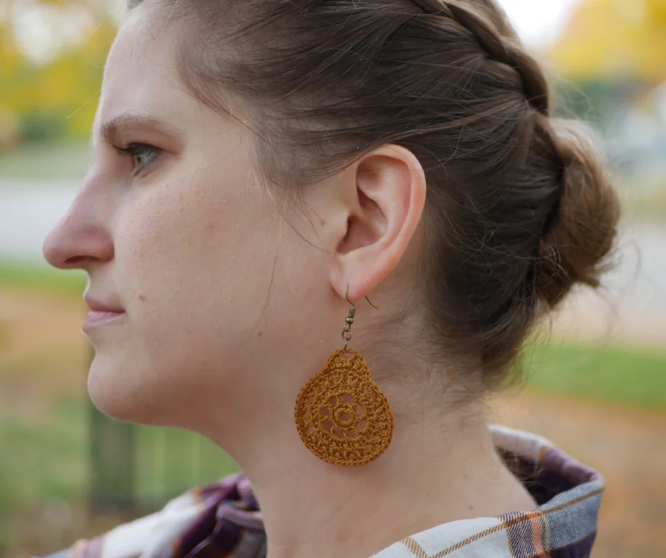 Image of a white woman in profile with light brown hair braided on the side and pulled up at the base of the neck. There is a yellow, circular crochet earring handing from her earlobe. 