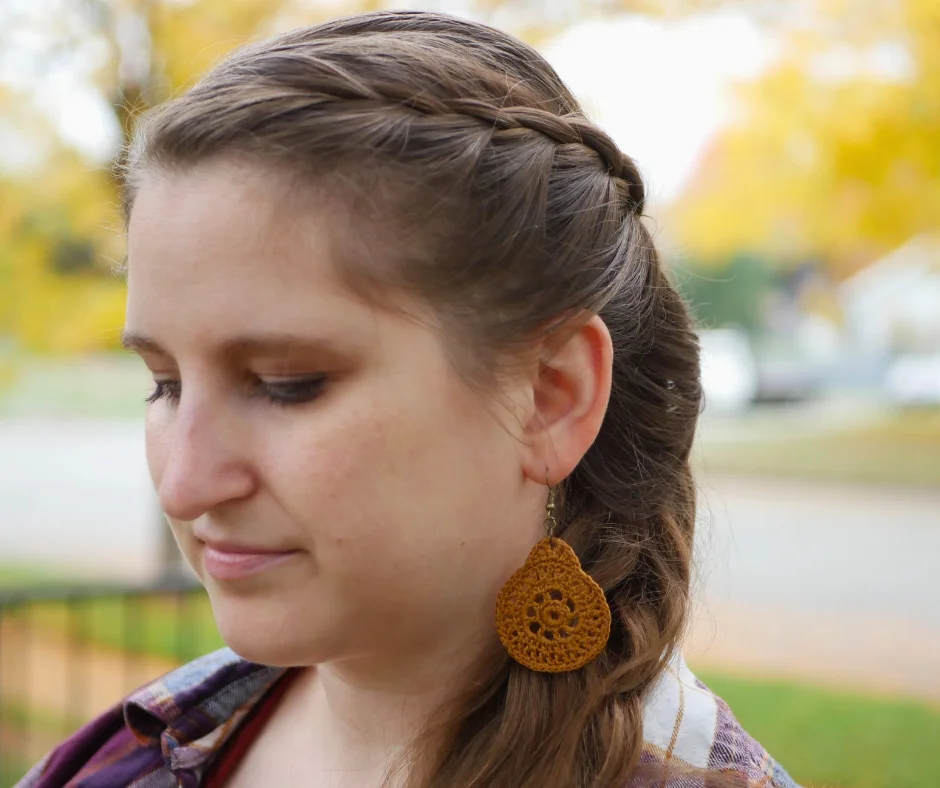 Image of a white woman wearing a flannel shirt outside in profile with her eyes downcast. A yellow equinox earring hands from her ear. 