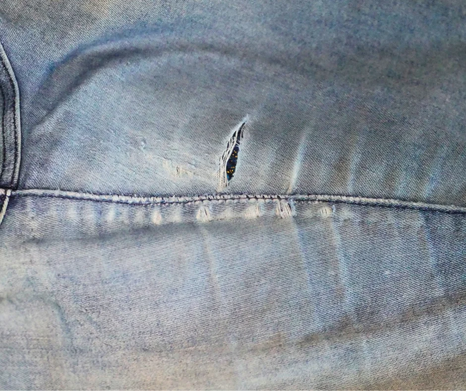 Image of the outside of of the inner thigh of a pair of jeans that is worn down and holey in places. Poking out of the hole is a dark blue fabric with gold specks. 