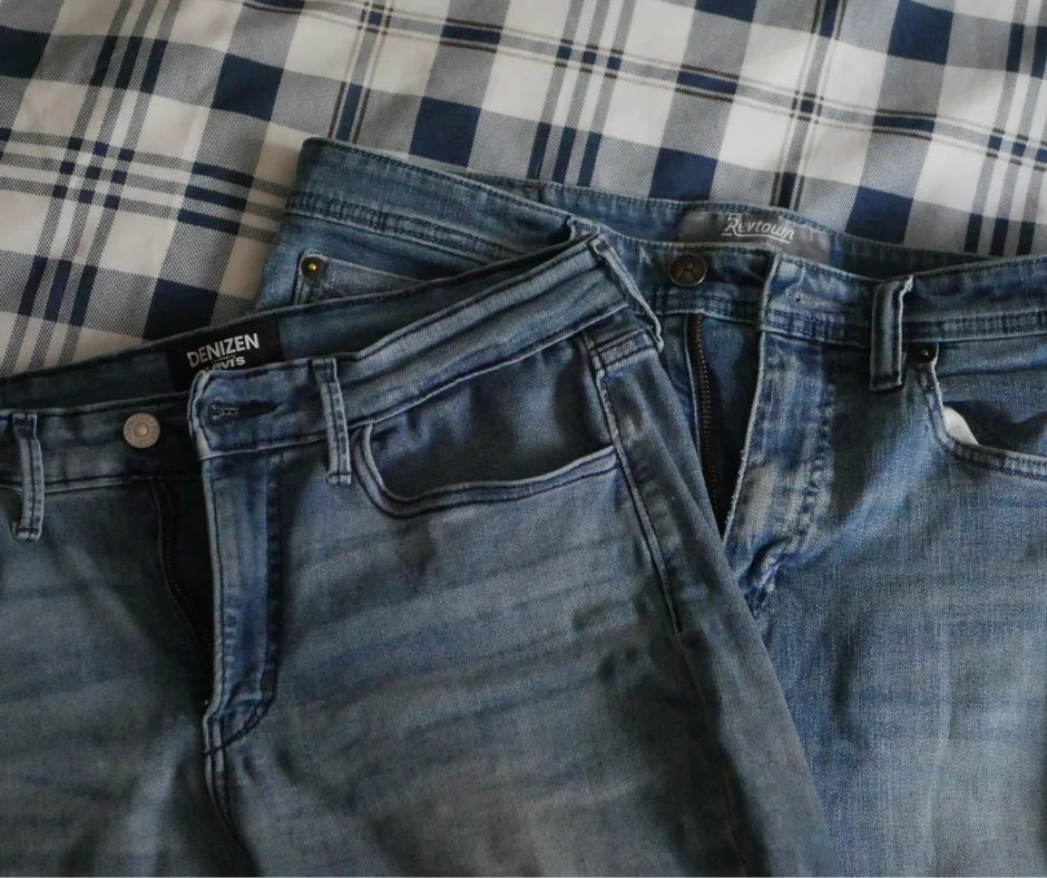 Image of the top front of two pairs of light blue denim jeans laying on a blue plaid background. 