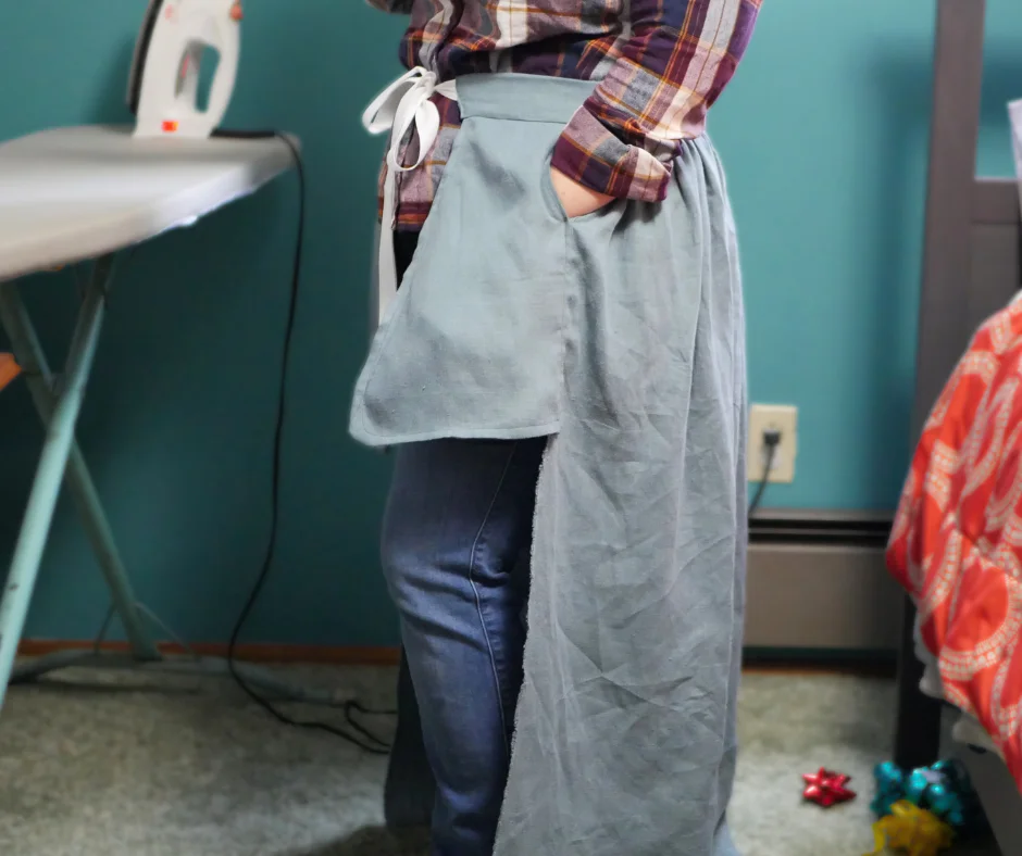 Image of a white person wearing jeans and a plaid shirt with the back panel of a blue linen split side skirt tied around their waist. 