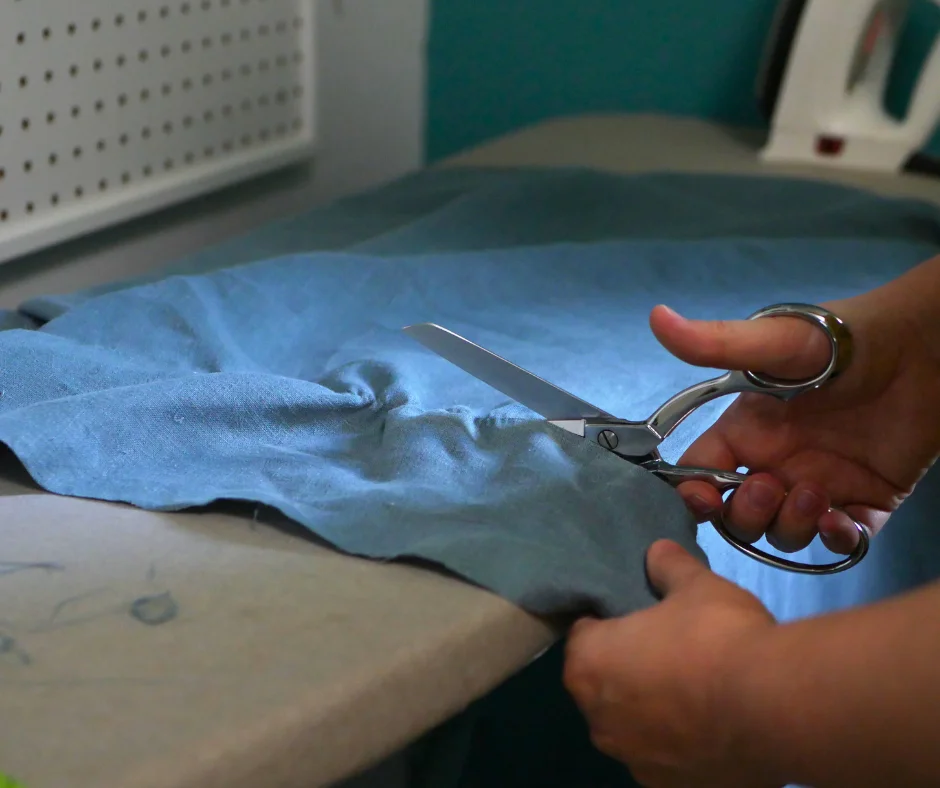Image of a pair of white hands holding steel fabric shears cutting blue linen fabric with a gather marking where to cut. 