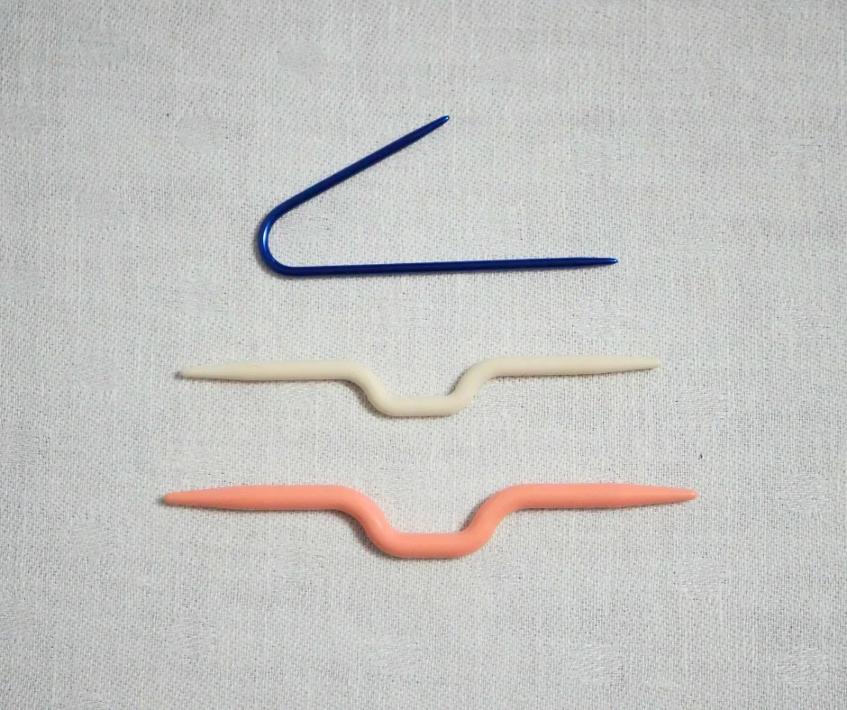 Image of three different cable needles. Top needle is blue metal in a U shape with one side longer than the other. Middle cable needle is white plastic, pointed on each end, with a bump in the middle. The bottom cable needle is salmon colored plastic, the same shape as the middle needle, but larger. 