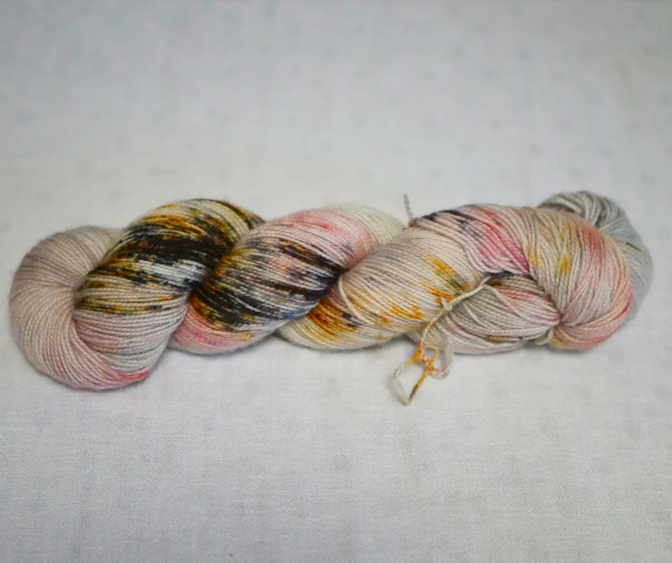 Image of a hank of yarn twisted up. Yarn is cream with browns, golds, and pinks over dyed. 