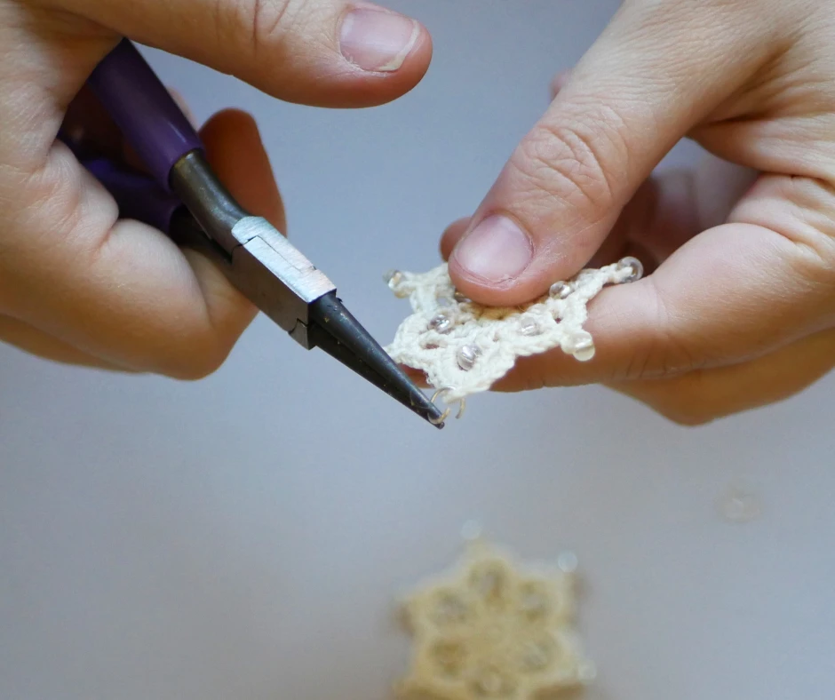 Image of a white pair of hands adding a jump ring to the top of a crochet snowflake with a pair of pliers. 