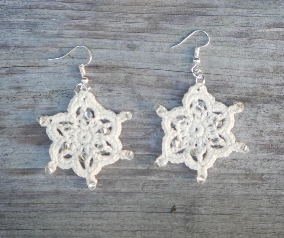 Image of two white crochet snowflake earrings with seed beads resting on a grey wood background. 