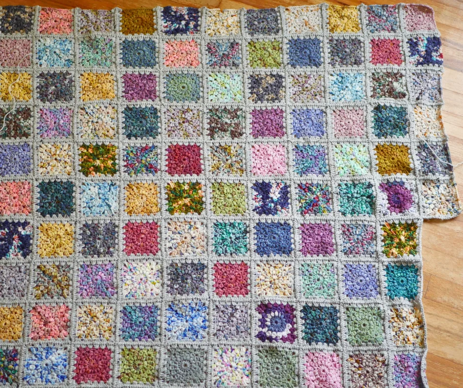 Image of a crochet granny square blanket with different colored squares laying on a wood floor. 
