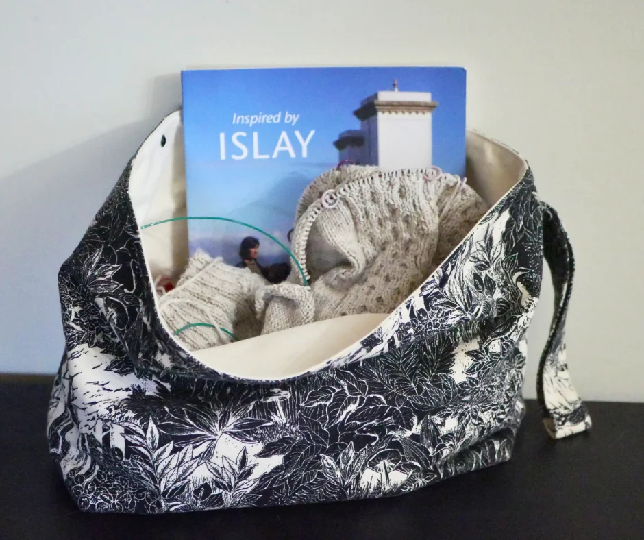 Image of a large black and white project bag with cream colored knitting and a knitting pattern book titled Inspired by Islay inside. 