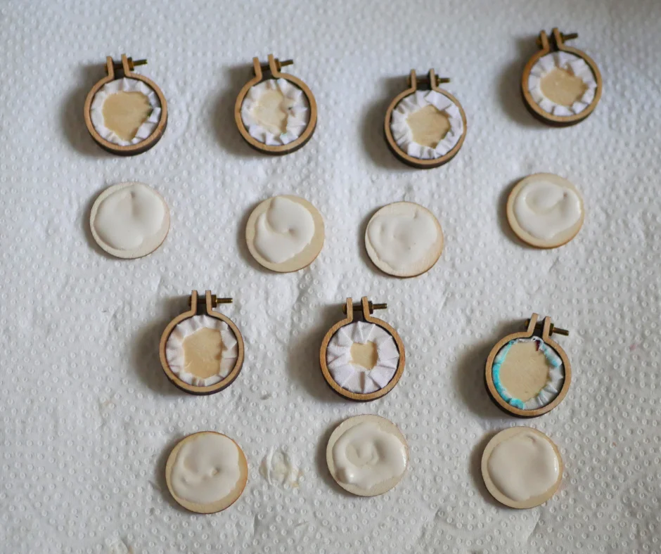 Image of the back of seven mini embroidery hoops with the screw added to the top of each. The back edges of fabric from the front embroidery are visible. There is a wood disc for each hoop with a generous amount of glue added, about to be placed on the the backs of the embroidery hoops. 