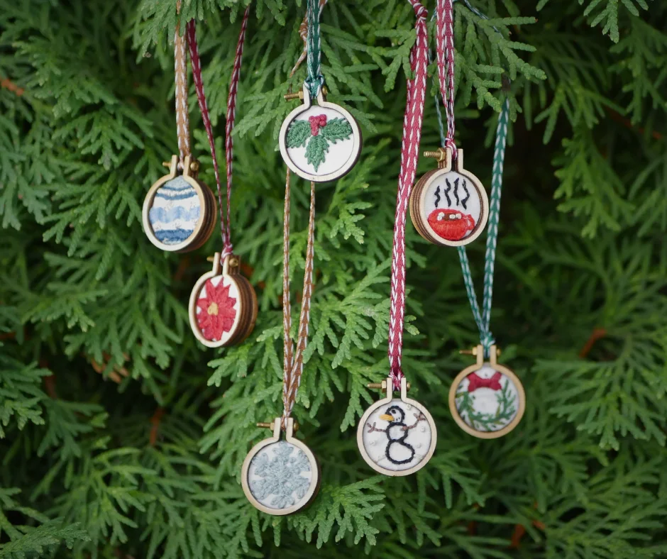 Image of seven mini Christmas embroidered ornaments hanging on some greenery. 