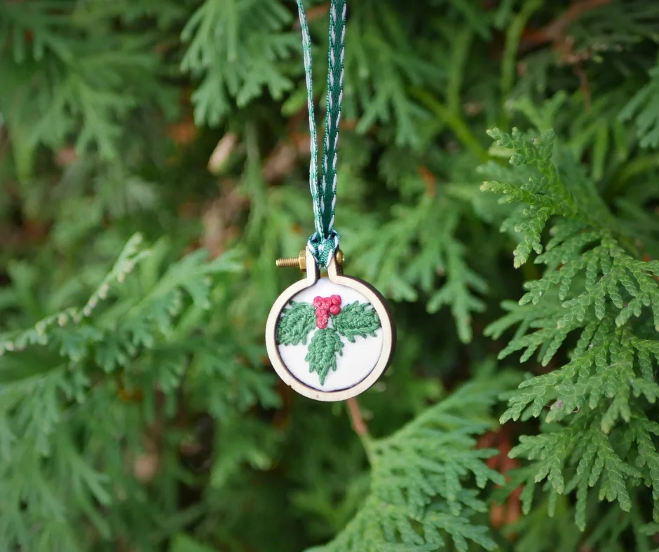 Image of an embroidered holly Christmas ornament in front of greenery. 